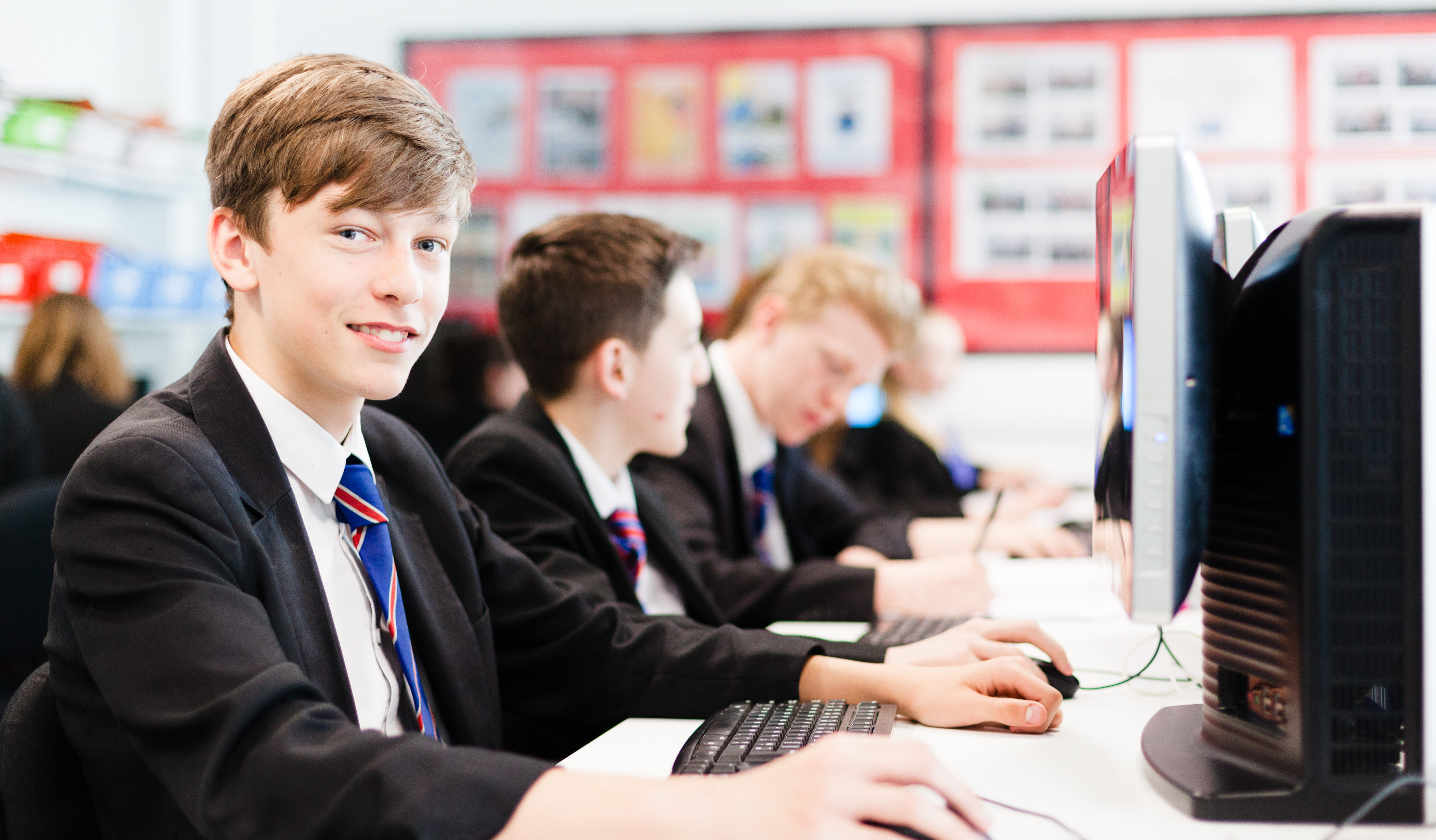 3 male students sitting at computers with the closest student looking and smiling at the camera